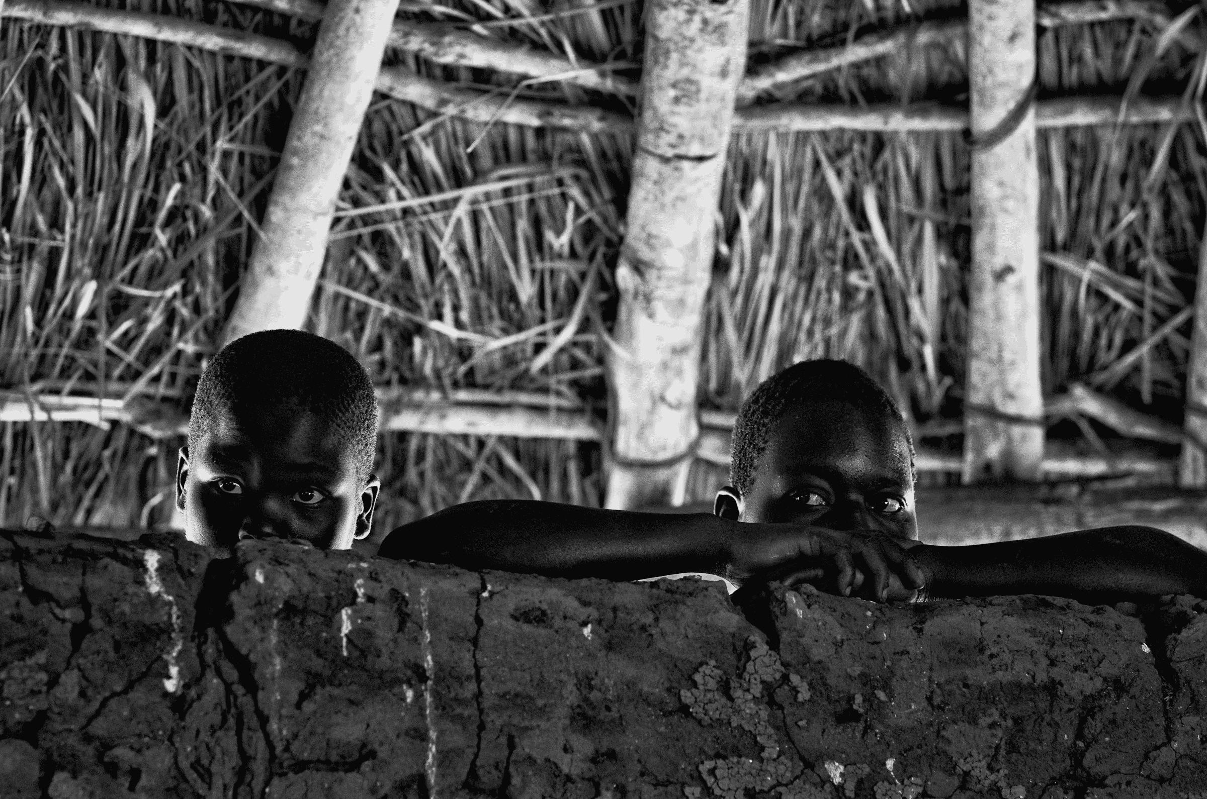 Black and white photograph of two young children in North Uganda peeking over a rough mud wall, looking directly at the camera, inside a traditional structure..