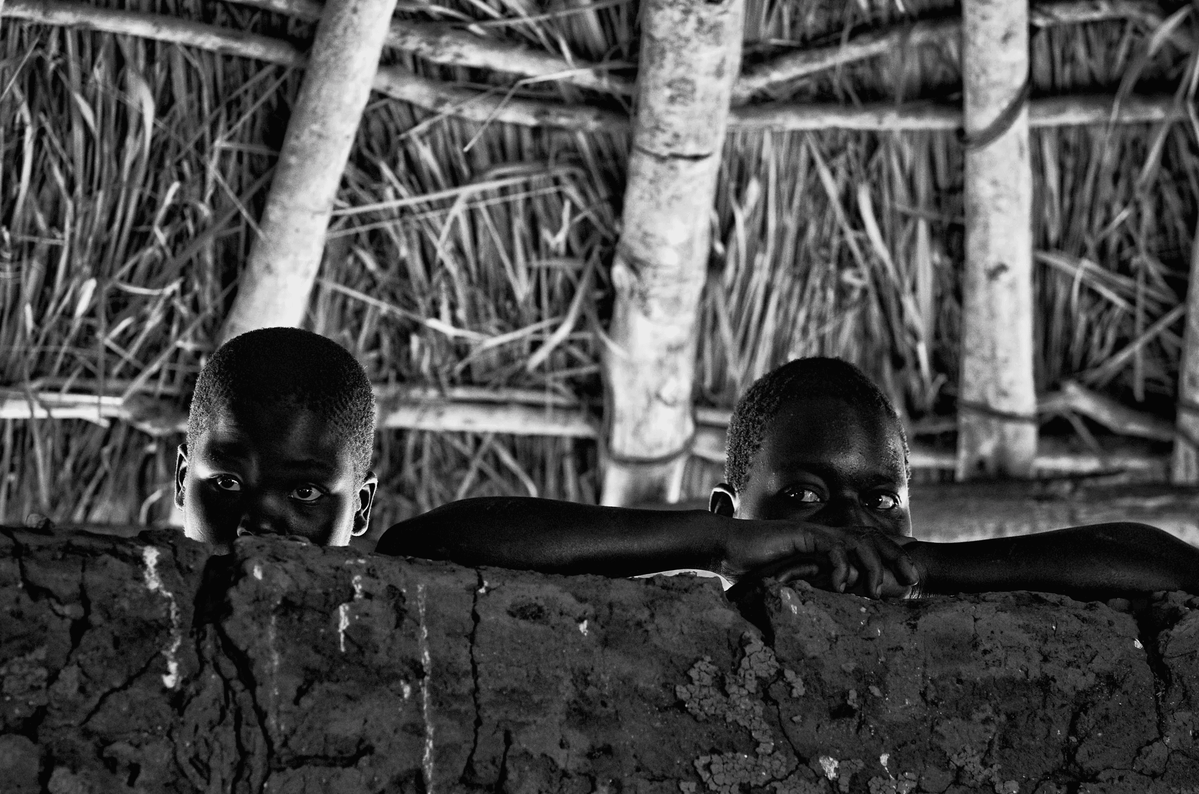 Black and white photograph of two young children in North Uganda peeking over a rough mud wall, looking directly at the camera, inside a traditional structure..