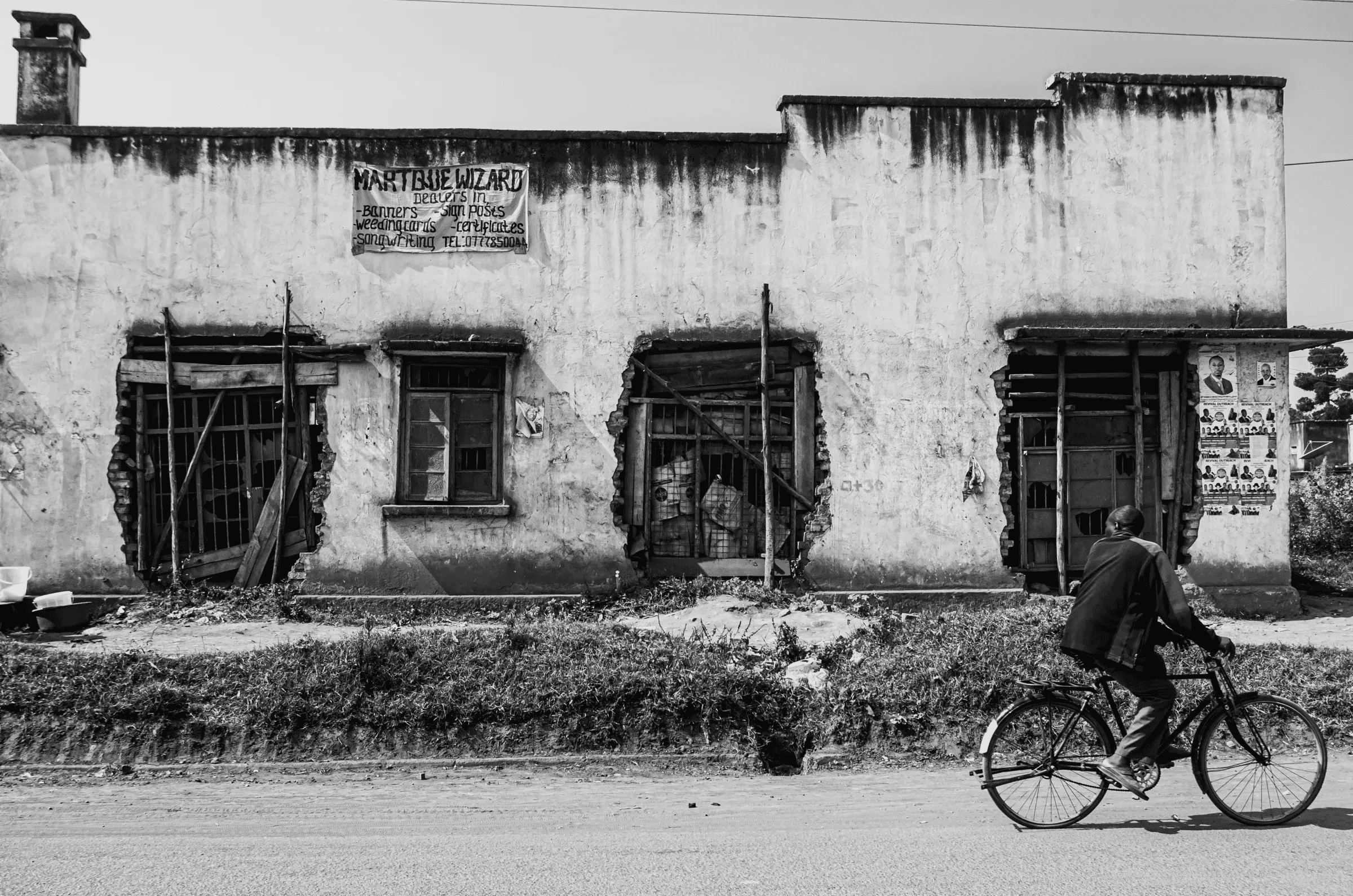 Black and white photograph of a person cycling past a weathered building with damaged windows on a roadside in Northern Uganda.