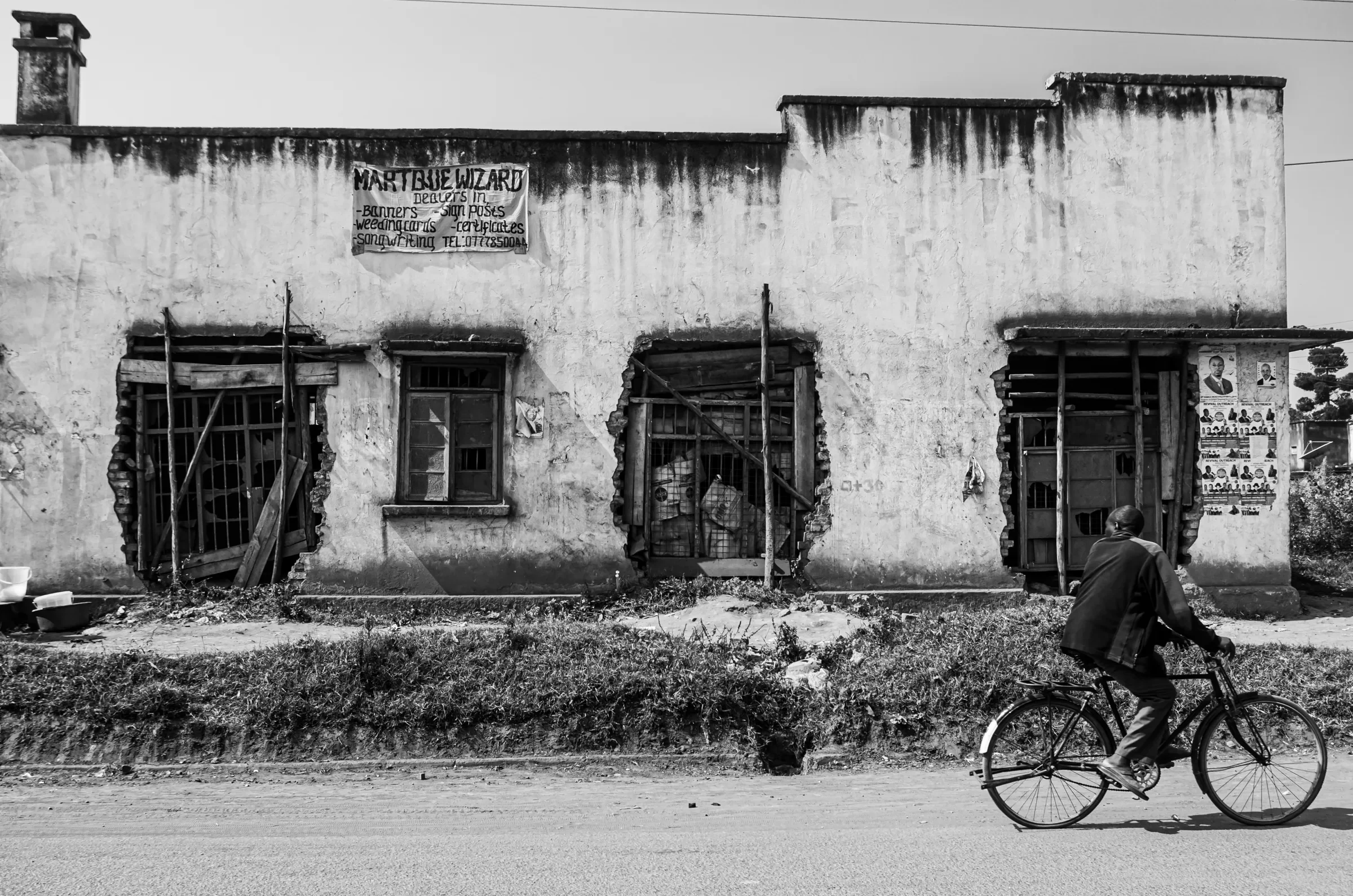 A person cycling past a weathered building with damaged windows on a roadside in Northern Uganda.