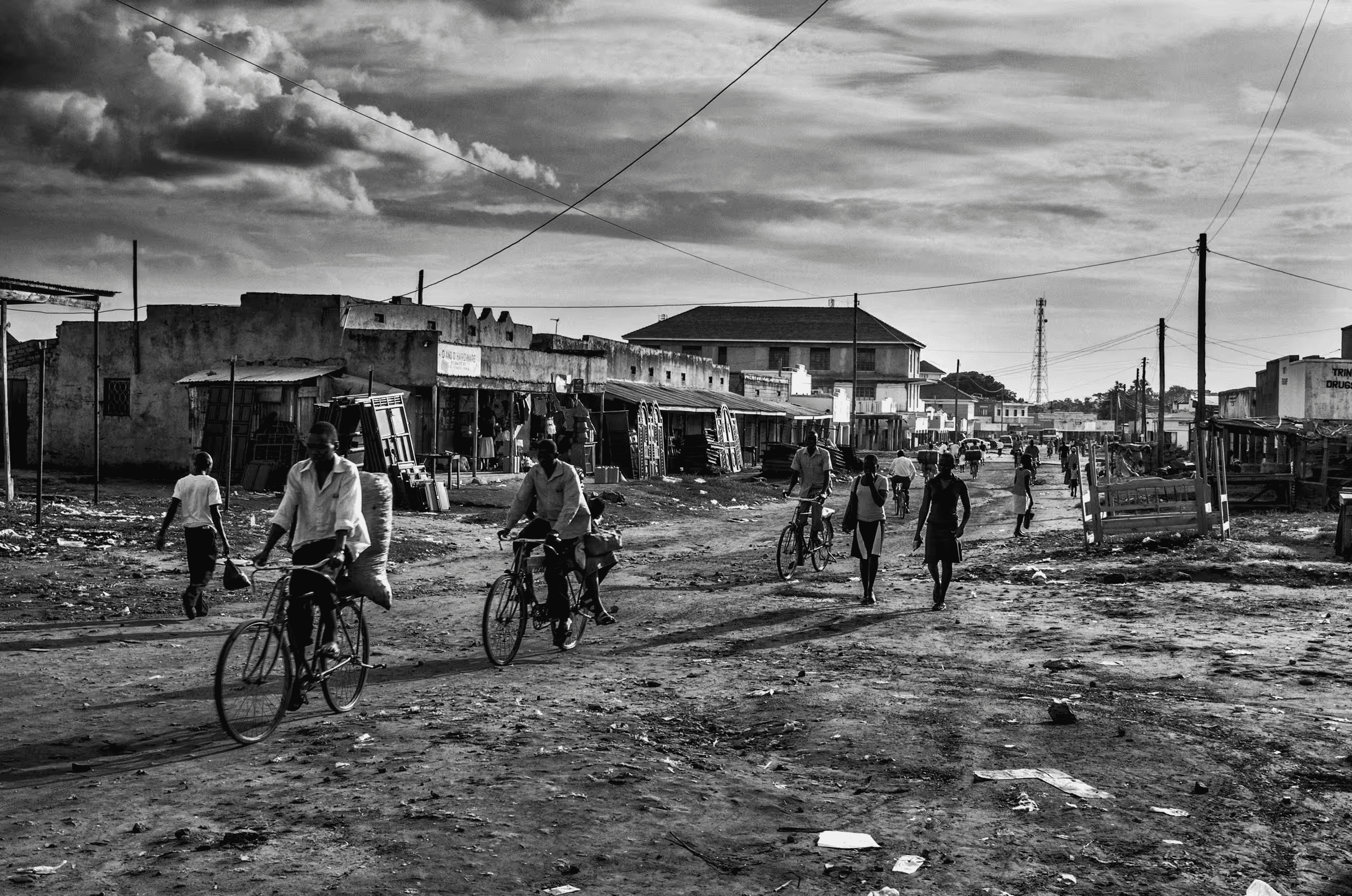 Black and white street scene photograph in a refugee settlement in Northern Uganda. People are walking and cycling on a dirt road lined with simple buildings and shops.