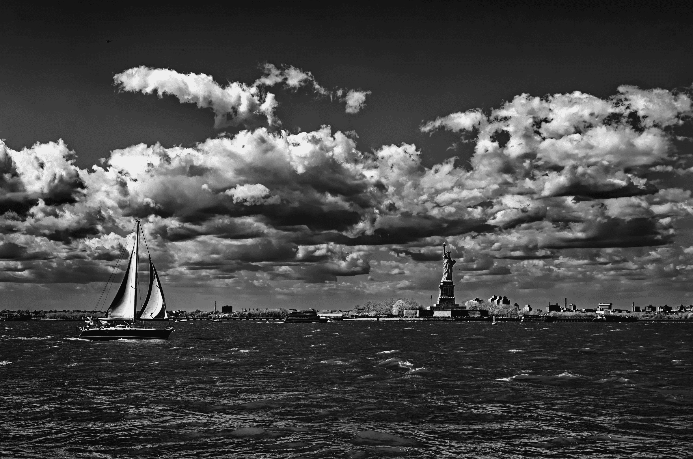 Dramatic black and white photograph of the Statue of Liberty and Lower Manhattan skyline viewed from New York Harbor, with a sailboat in the foreground under a cloudy sky.