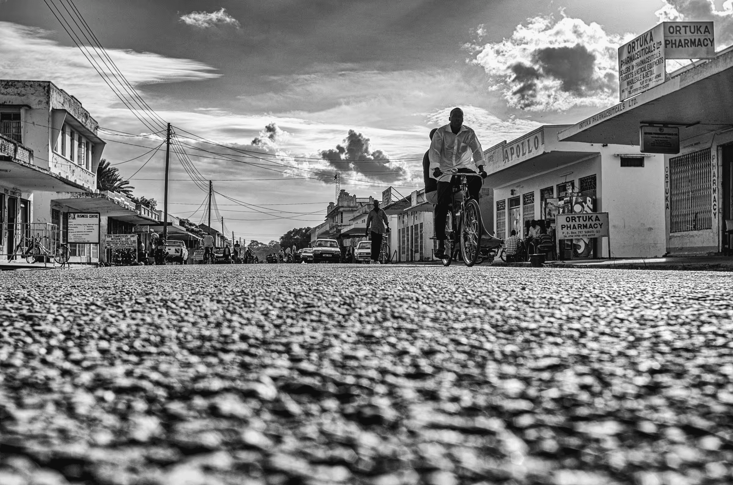 Black and white street scene photograph in Ortuka, Northern Uganda. People are walking and cycling on a dirt road.