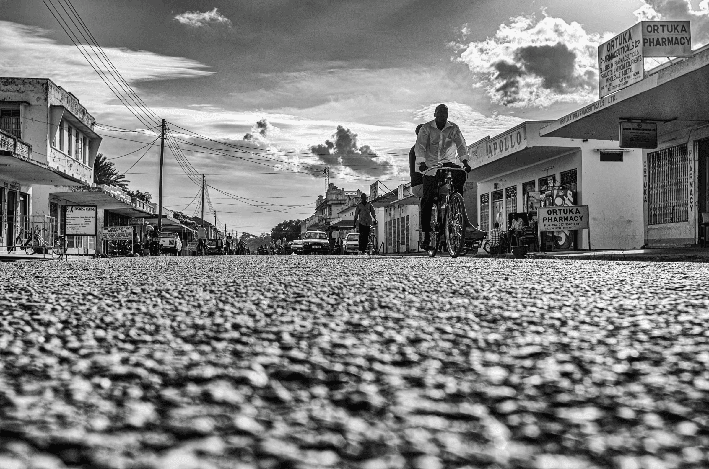 Black and white street scene photograph in Ortuka, Northern Uganda. People are walking and cycling on a dirt road.
