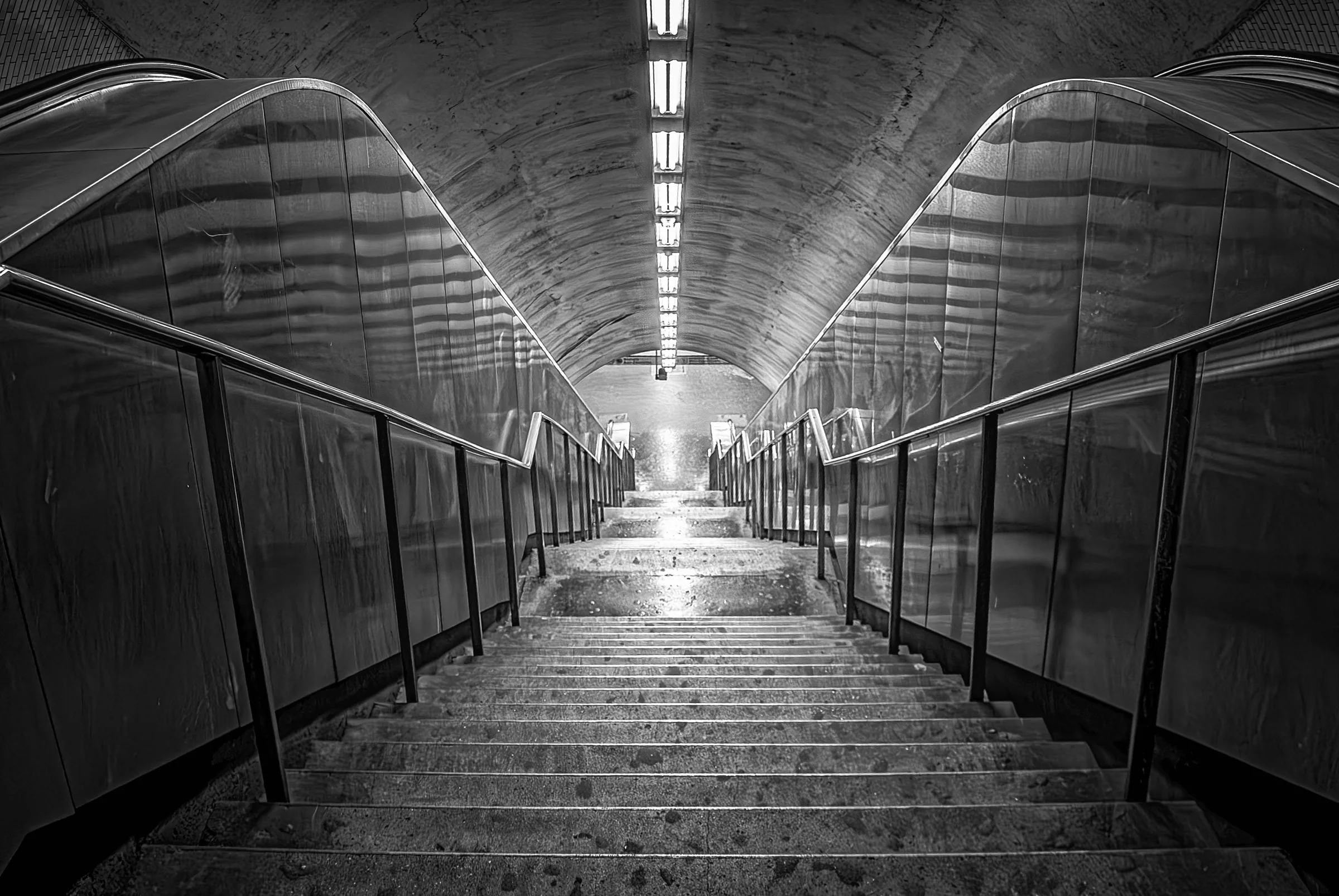 Black and white photograph of a long, empty staircase descending into a subway station. The image emphasizes the symmetry and lines of the staircase, handrails, and the curved, tiled ceiling.