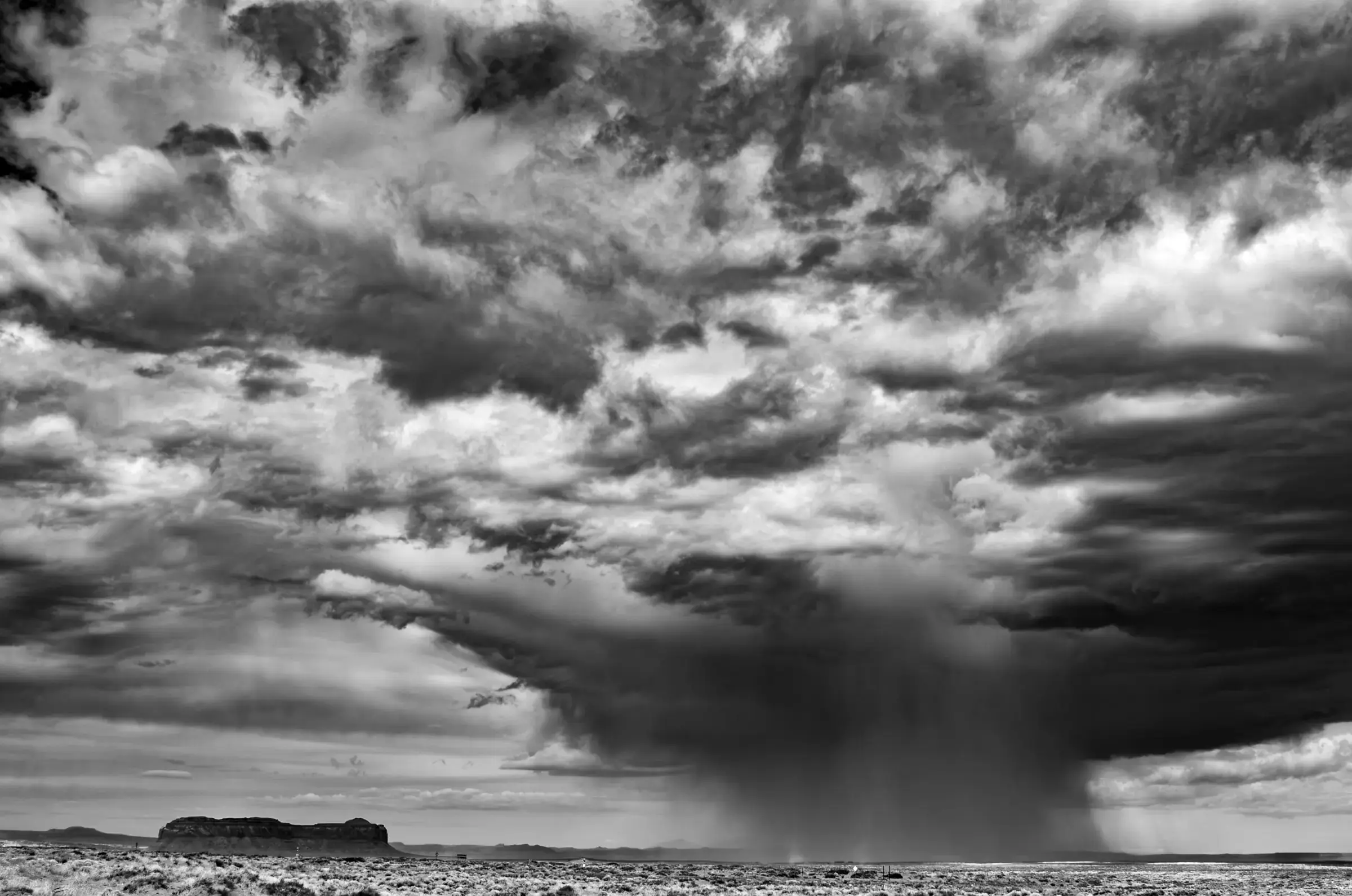 Rainfall, High Desert Black and white photograph of a dramatic sky over a flat desert landscape, with a heavy rain shaft falling from dark clouds near a distant mesa.