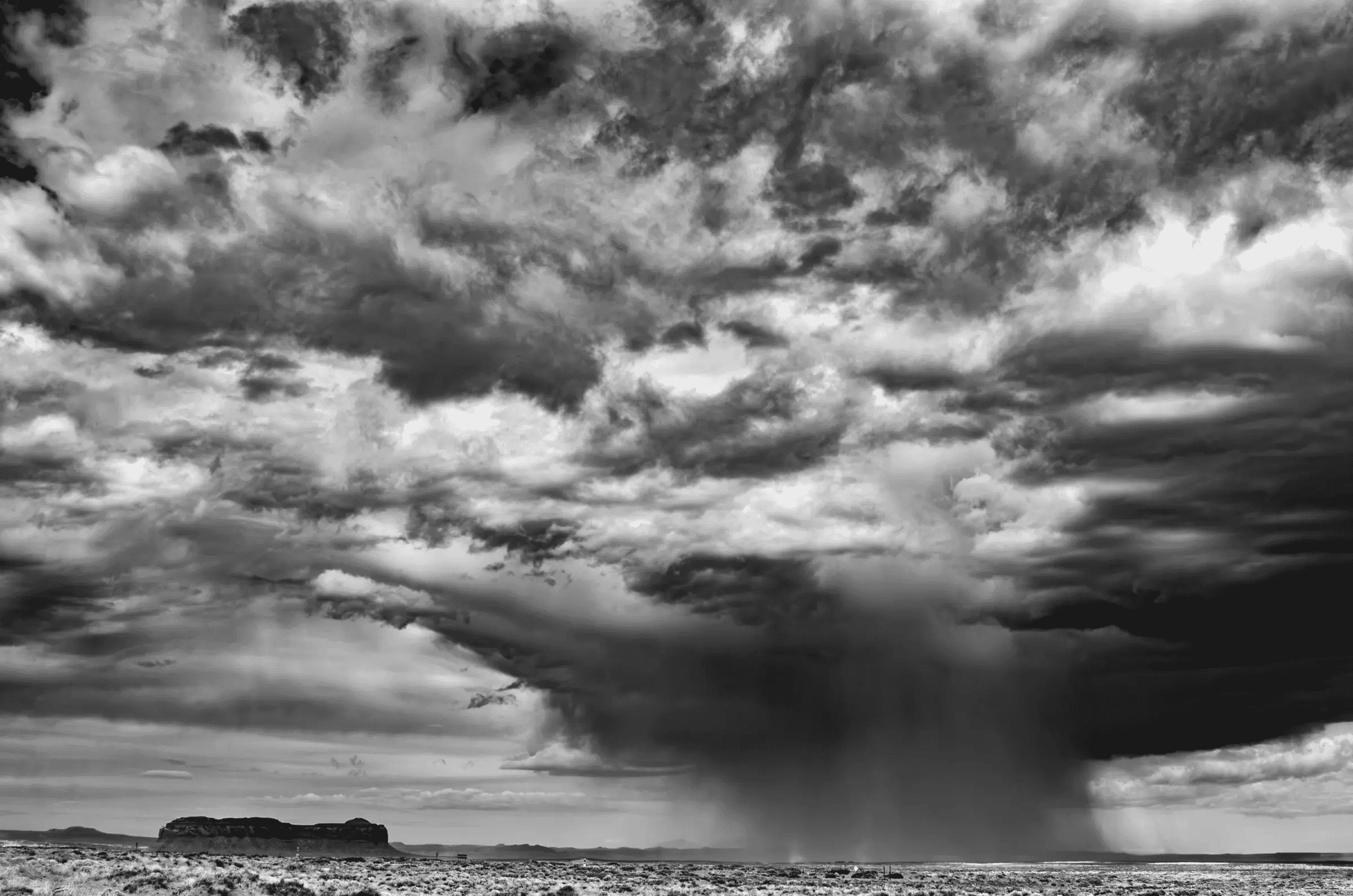 Rainfall, High Desert Black and white photograph of a dramatic sky over a flat desert landscape, with a heavy rain shaft falling from dark clouds near a distant mesa.