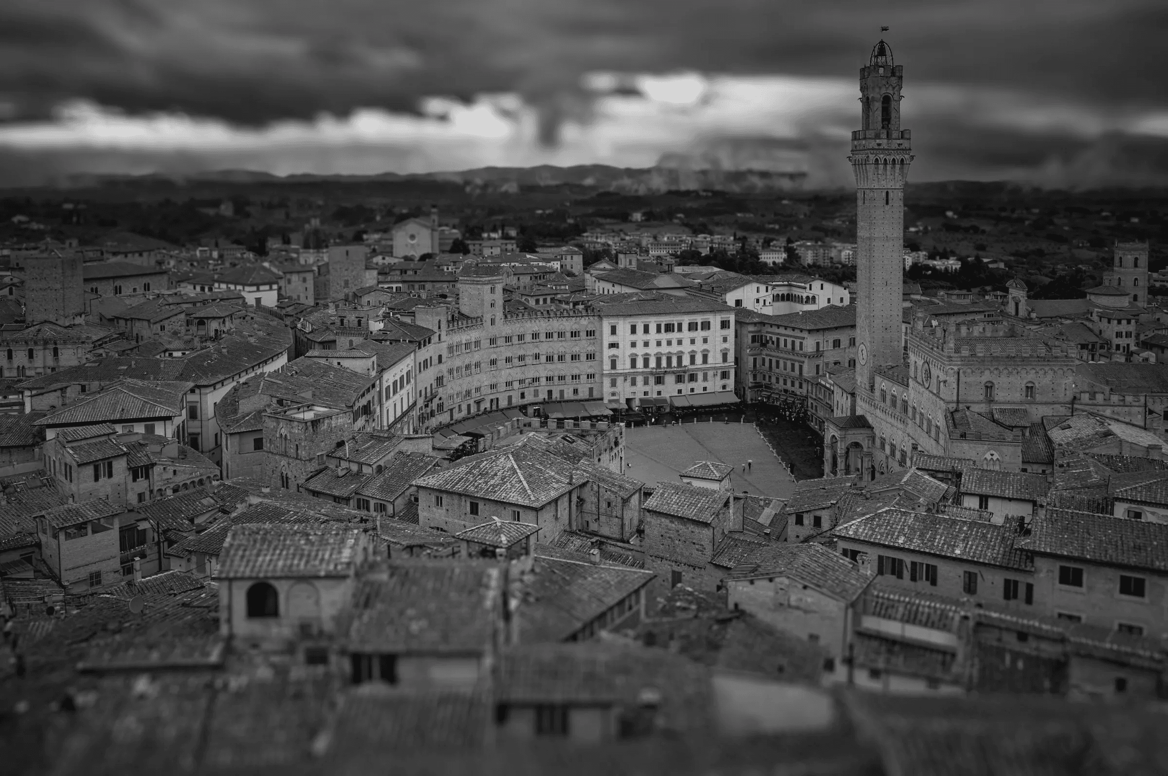 Black and white photograph taken from a high angle overlooking the Piazza del Campo in Siena, Italy, showing the distinctive curved piazza, surrounding buildings, and the Torre del Mangia under a dramatic sky.