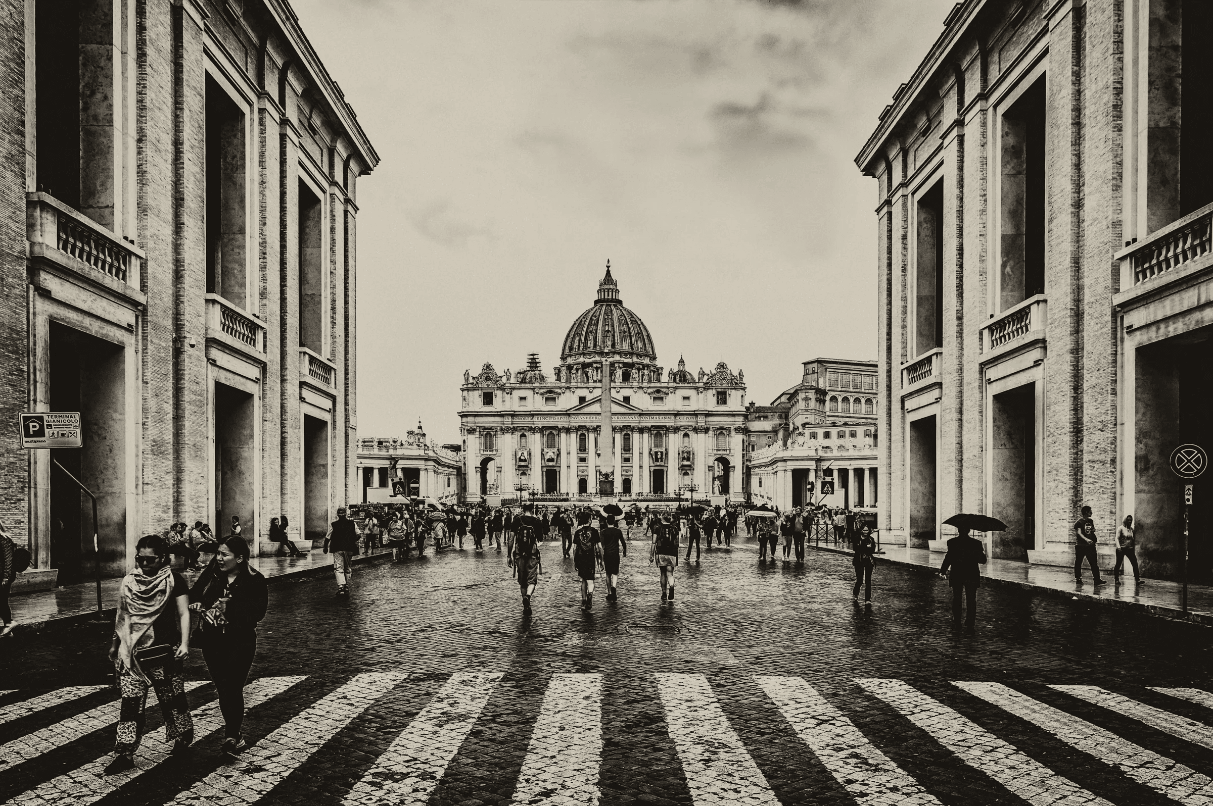 Monochrome photograph looking down the Via della Conciliazione towards St. Peter's Basilica and Square in Vatican City, with numerous pedestrians walking on the cobblestone street.