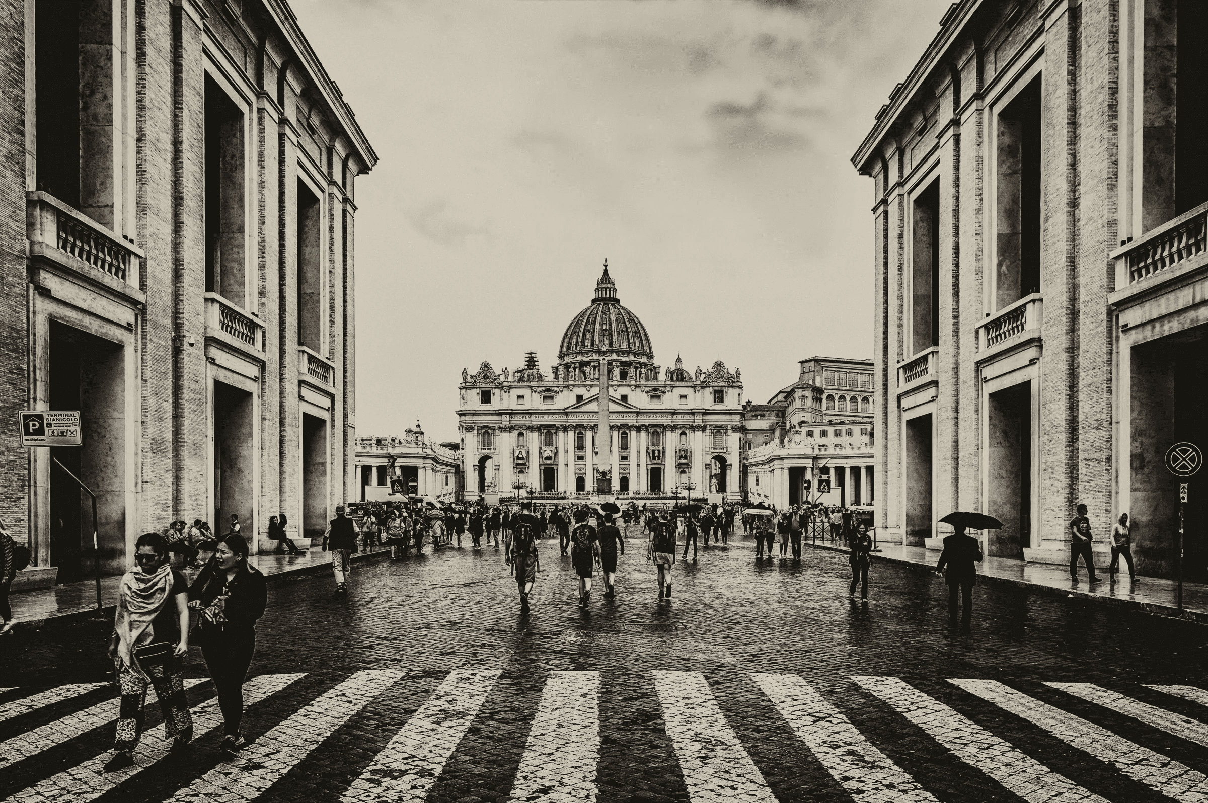 Monochrome photograph looking down the Via della Conciliazione towards St. Peter's Basilica and Square in Vatican City, with numerous pedestrians walking on the cobblestone street.