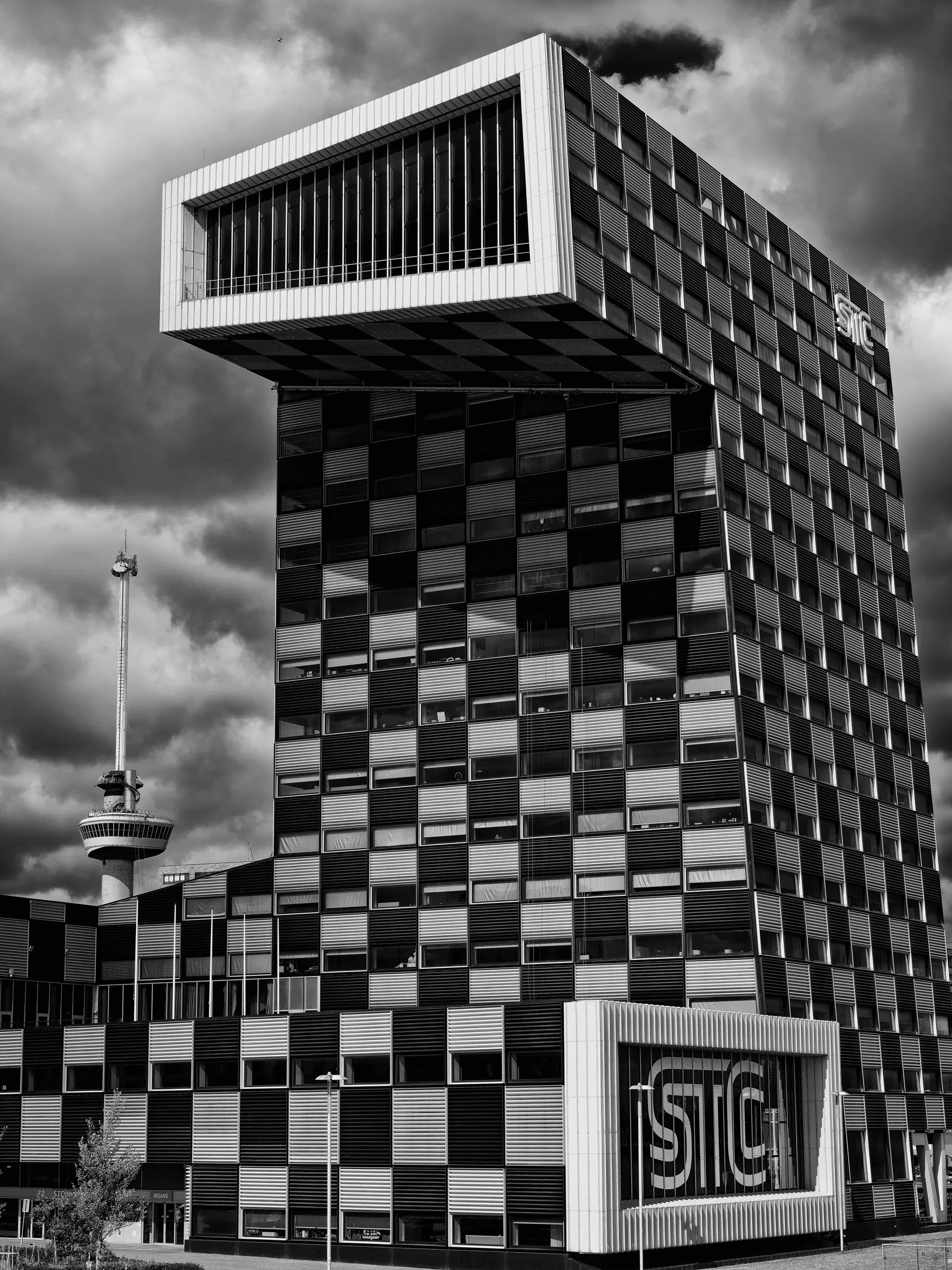 Black and white photograph of the STC building in Rotterdam with its bold geometric design, set against a moody sky and the Euromast tower in the background.