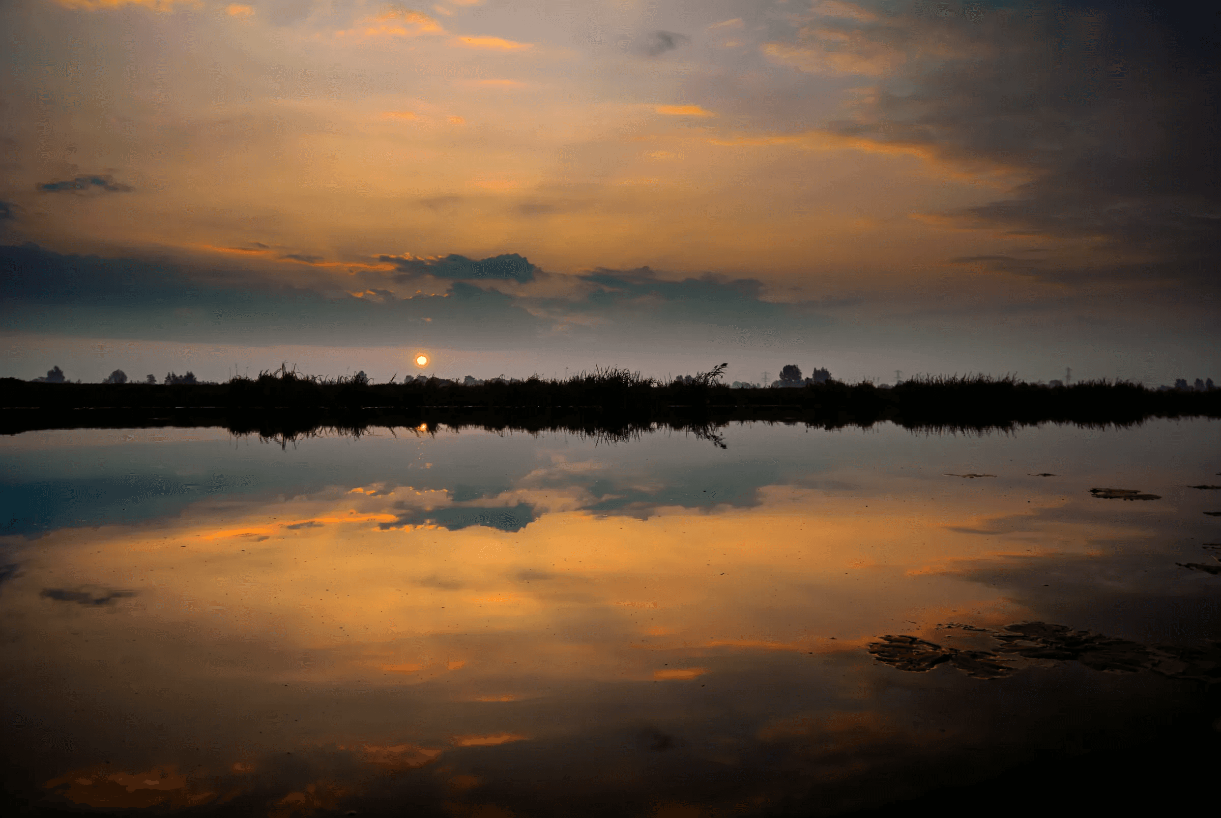 Vibrant sunset sky with orange and dark clouds reflected in calm water.