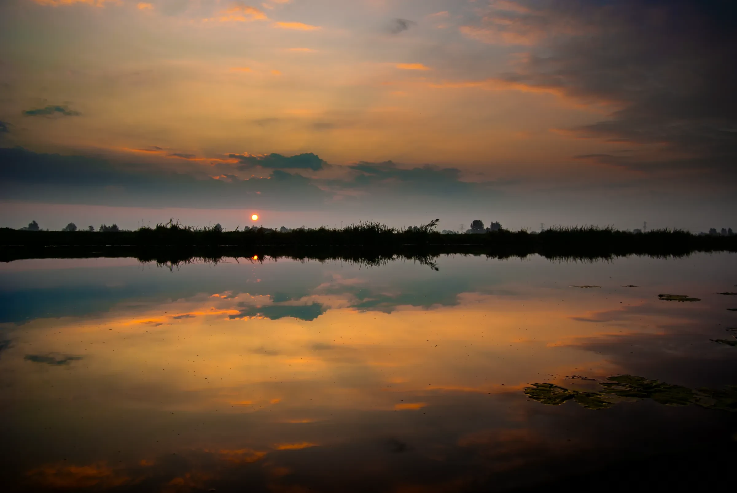 Vibrant sunset sky with orange and dark clouds reflected in calm water.