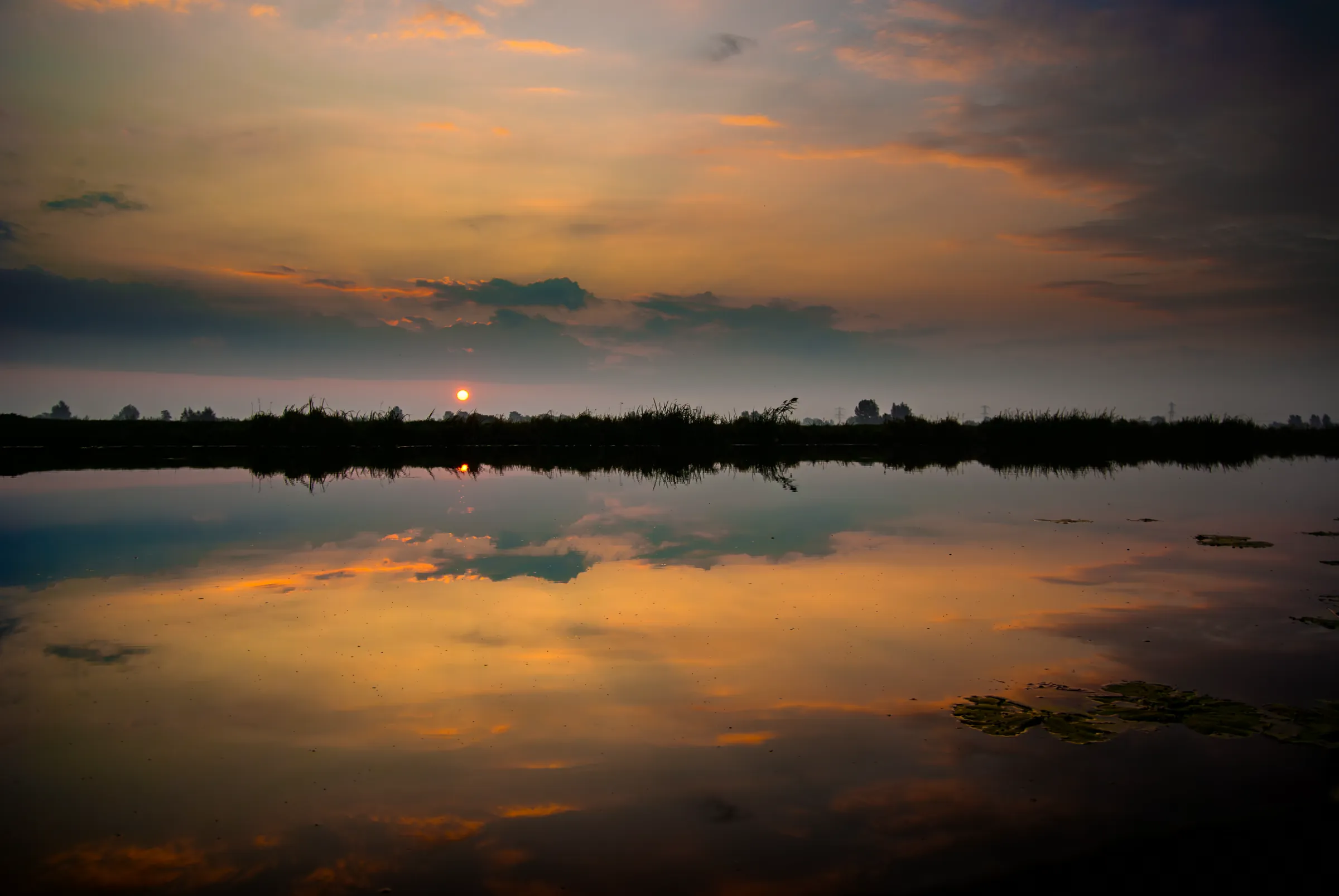 Vibrant sunset sky with orange and dark clouds reflected in calm water.