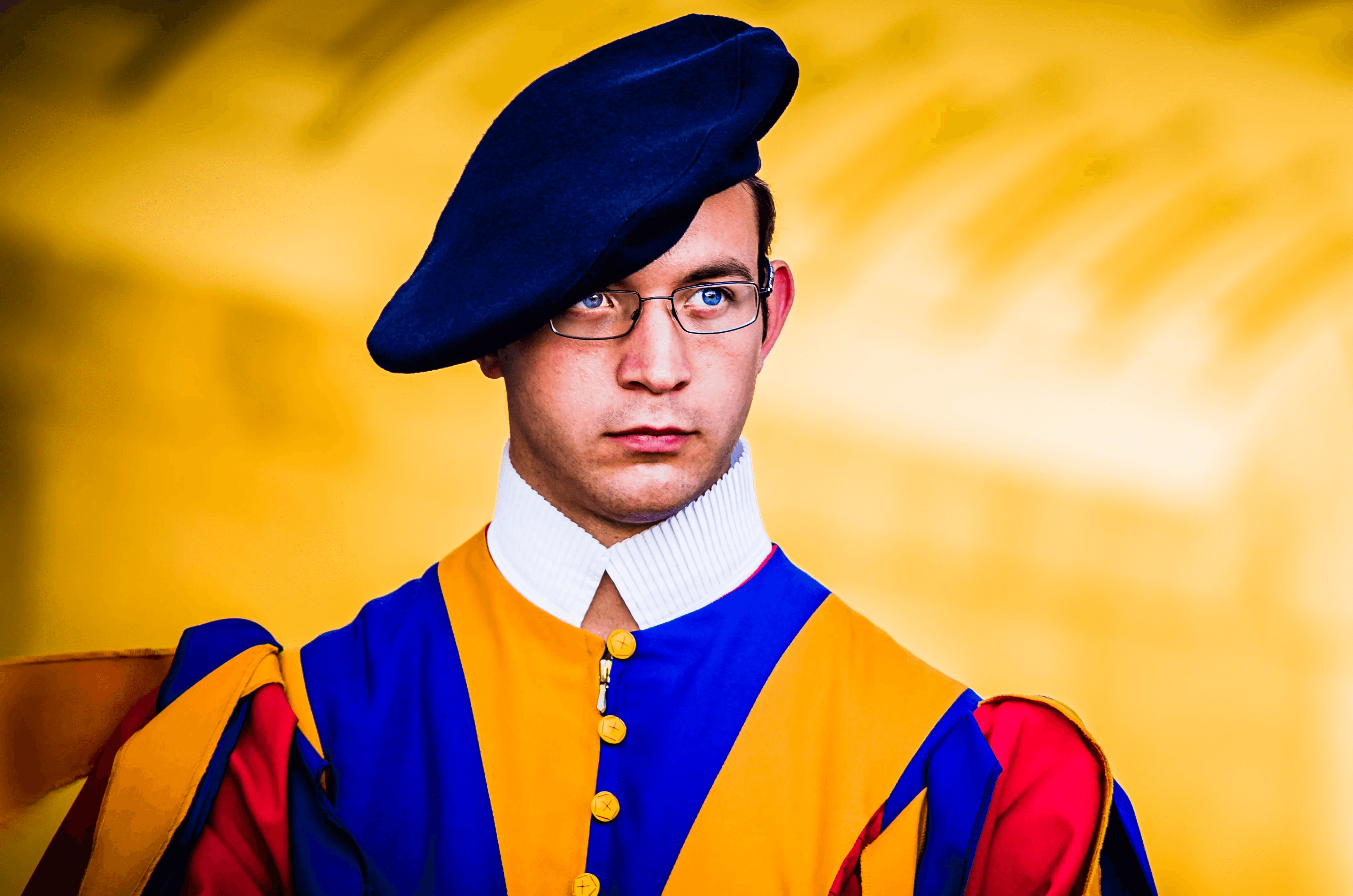 Close-up color portrait of a Swiss Guard at the Vatican. He wears the traditional colorful blue, yellow, and red Renaissance-style uniform, a dark blue beret, and glasses.