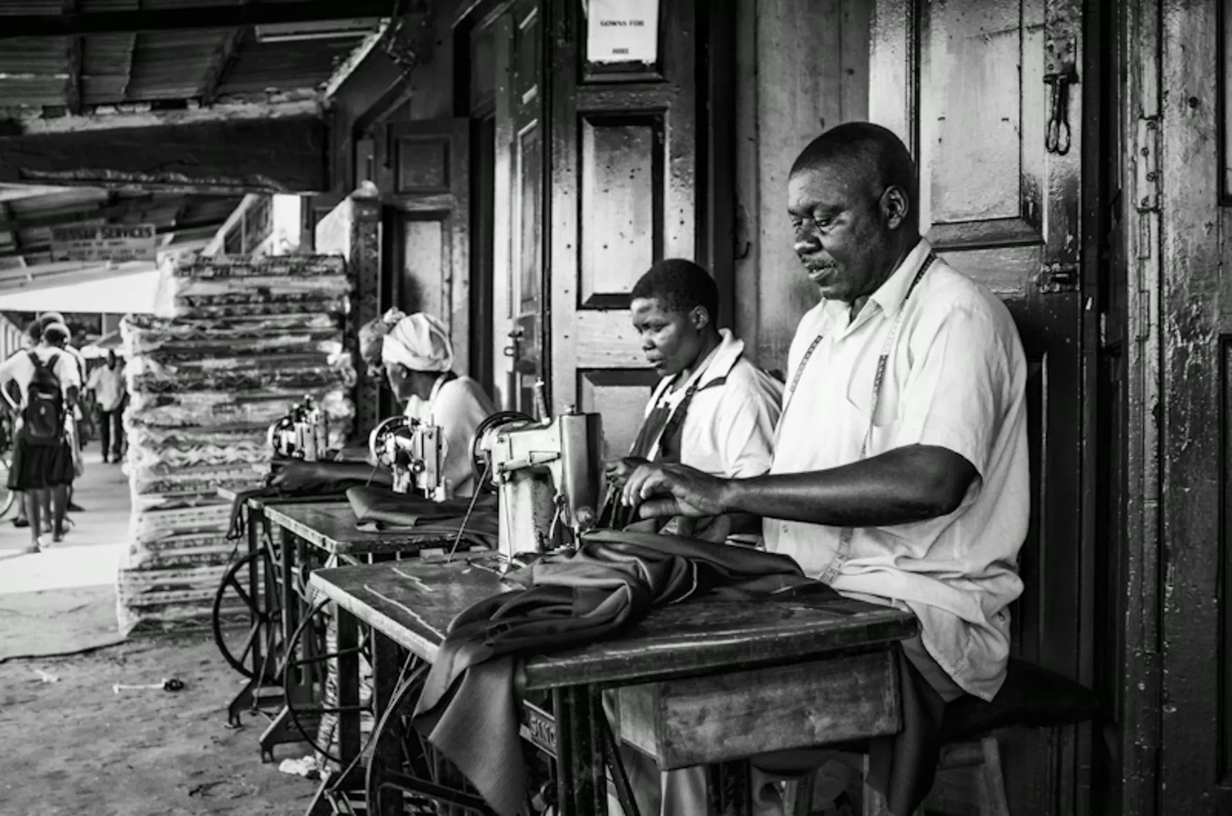 Black-and-white photograph of tailors working at sewing machines outdoors.