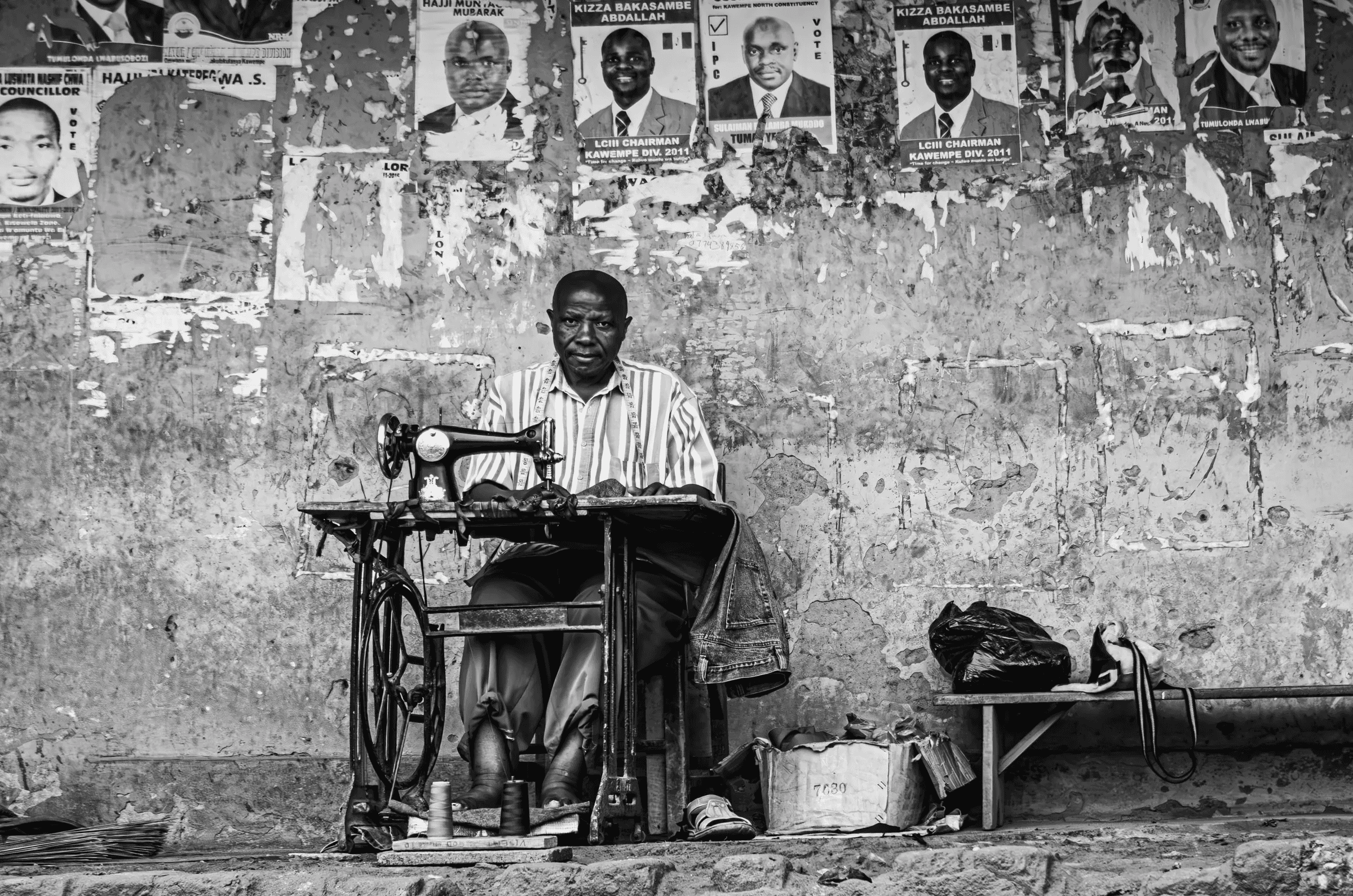 Black and white portrait of a man sitting at a treadle sewing machine outdoors in Uganda, looking at the camera, positioned against a weathered wall covered in torn political posters.