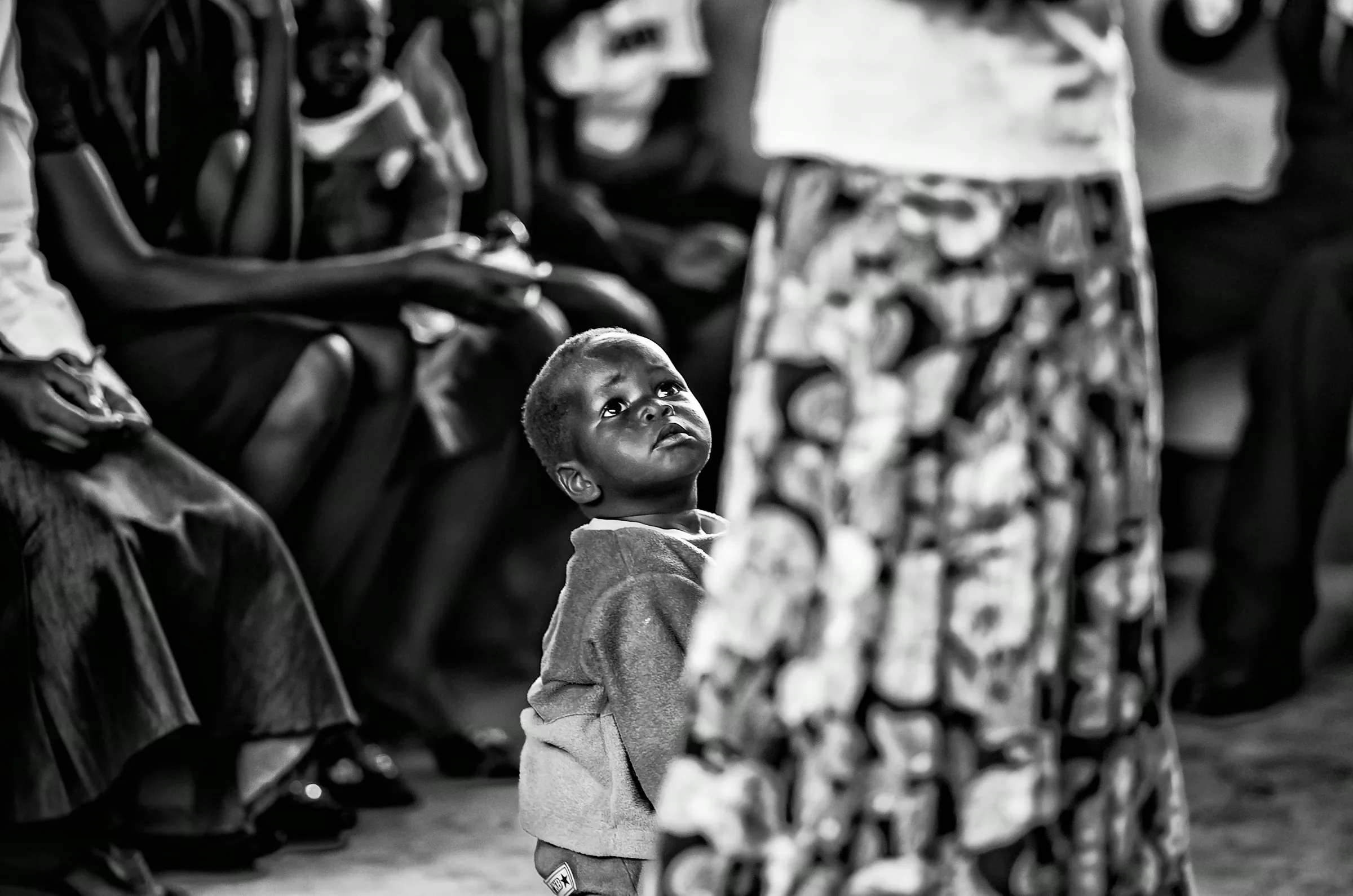 Black and white photograph focusing on a small Ugandan child looking upwards, partially obscured by an adult figure in the foreground.