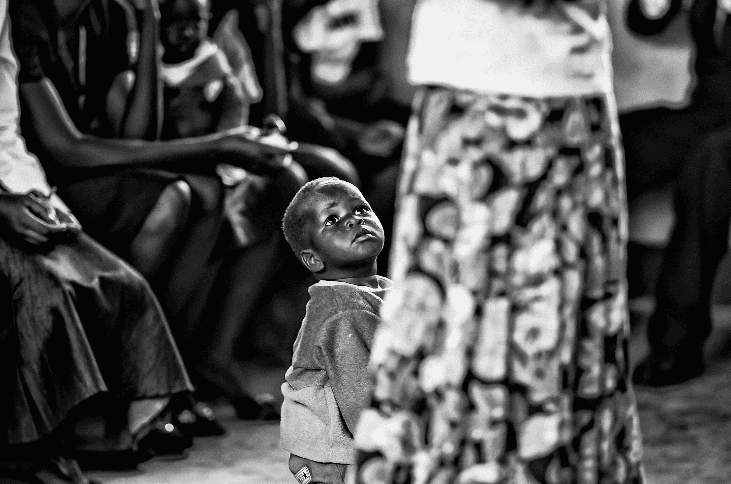 Black and white photograph focusing on a small Ugandan child looking upwards, partially obscured by an adult figure in the foreground.
