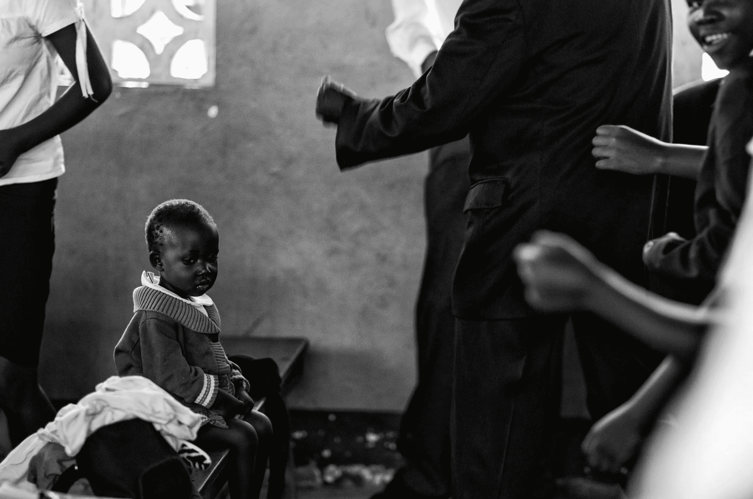 Black and white photograph of a small Ugandan child sitting quietly on a bench, observing activity in the background where other people are gathered indoors.