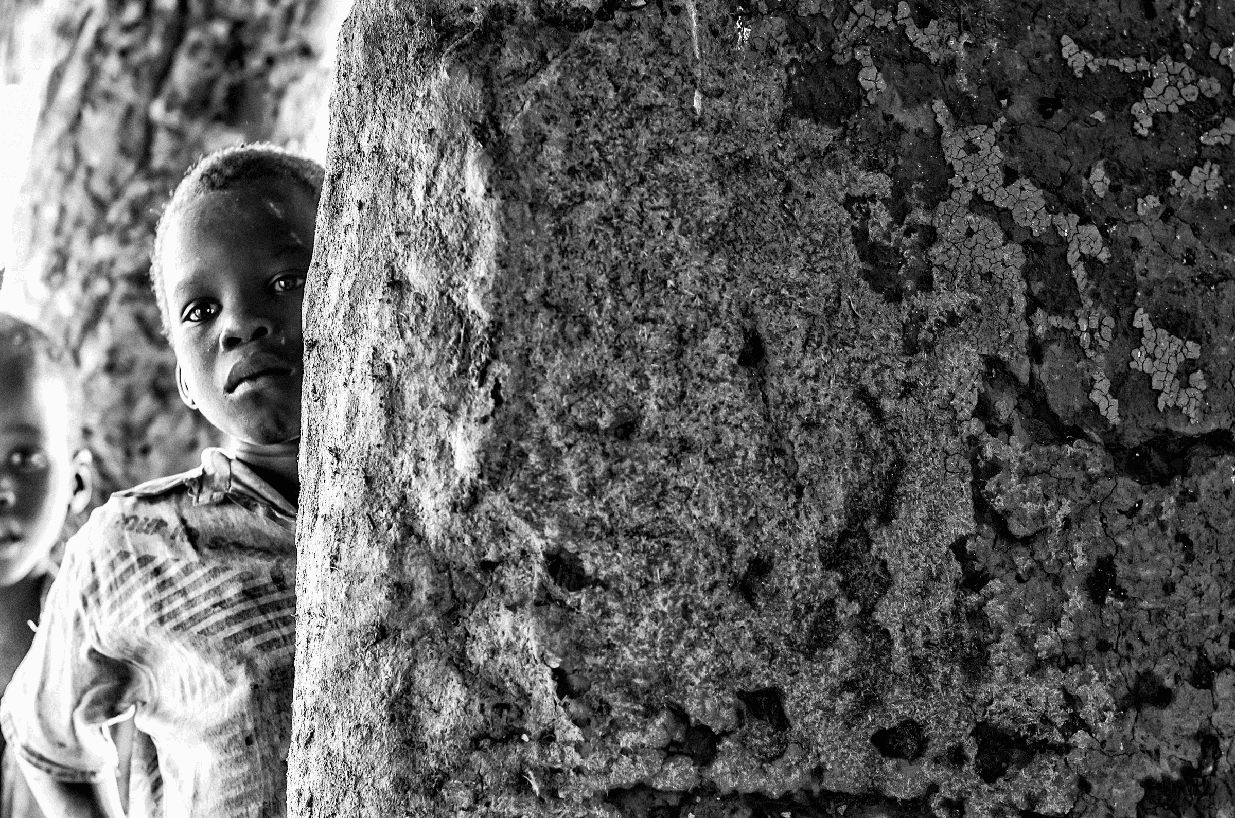 Peeking from Behind A young Ugandan child peeking around a rough, textured pillar or wall, looking towards the camera.