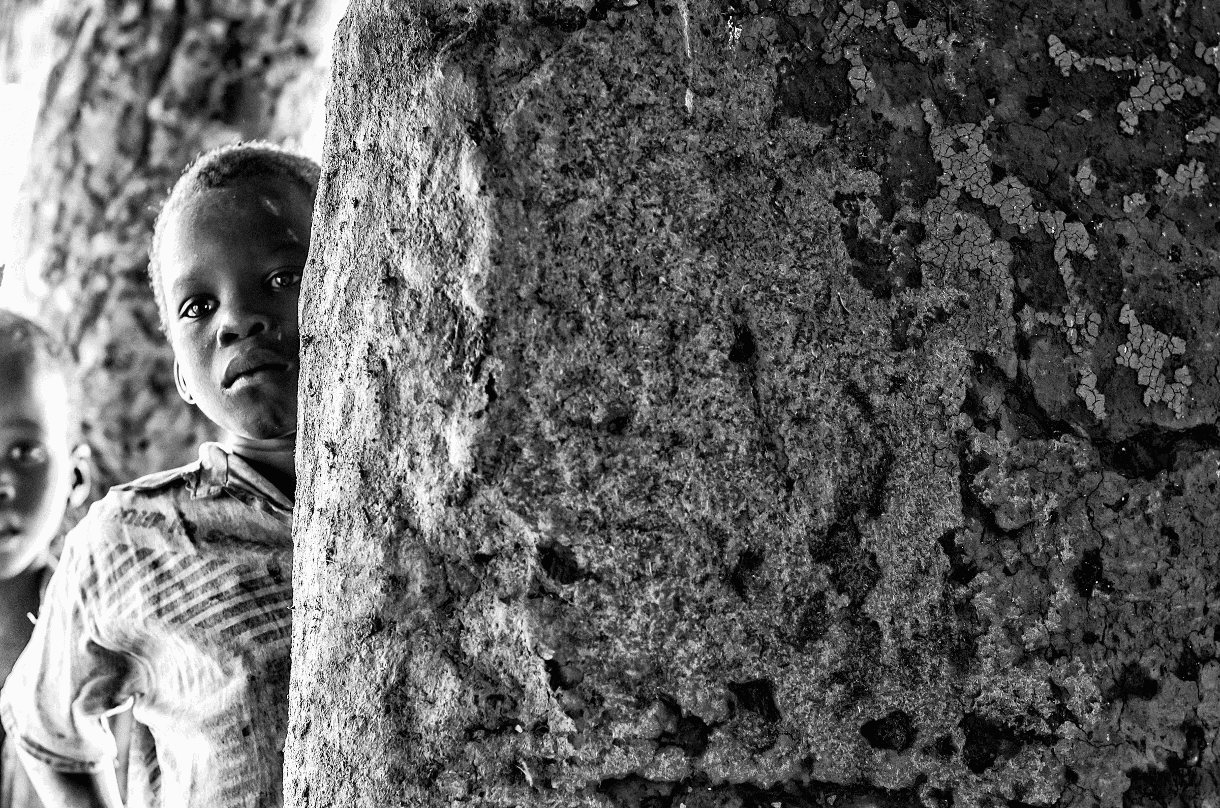 Black and white photograph of a young Ugandan child peeking around a rough, textured pillar or wall, looking towards the camera.