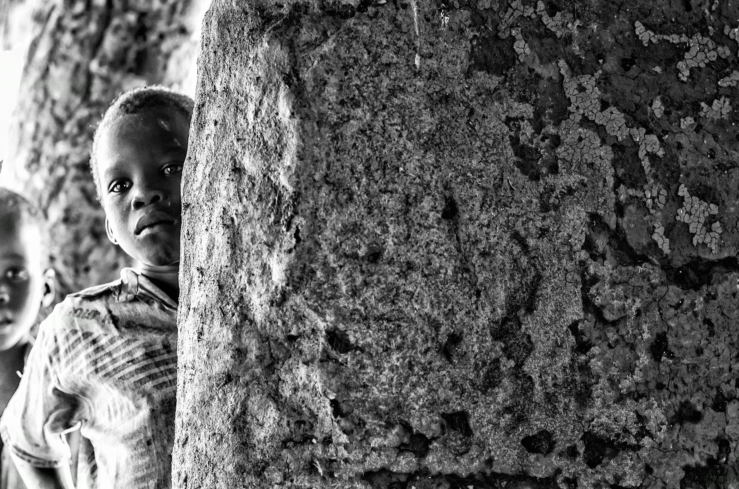 A young Ugandan child peeking around a rough, textured pillar or wall, looking towards the camera.