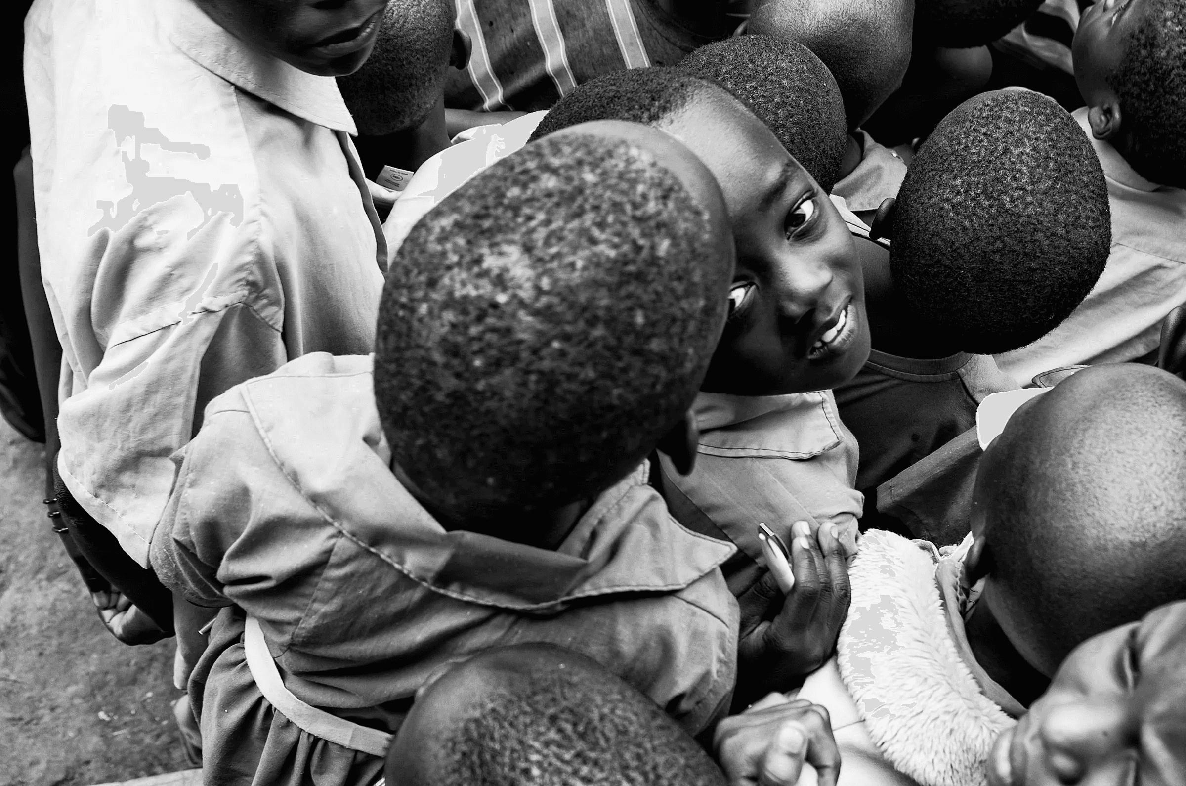 Black and white photograph taken from a high angle, looking down into a group of Ugandan children, one of whom looks directly up at the camera.