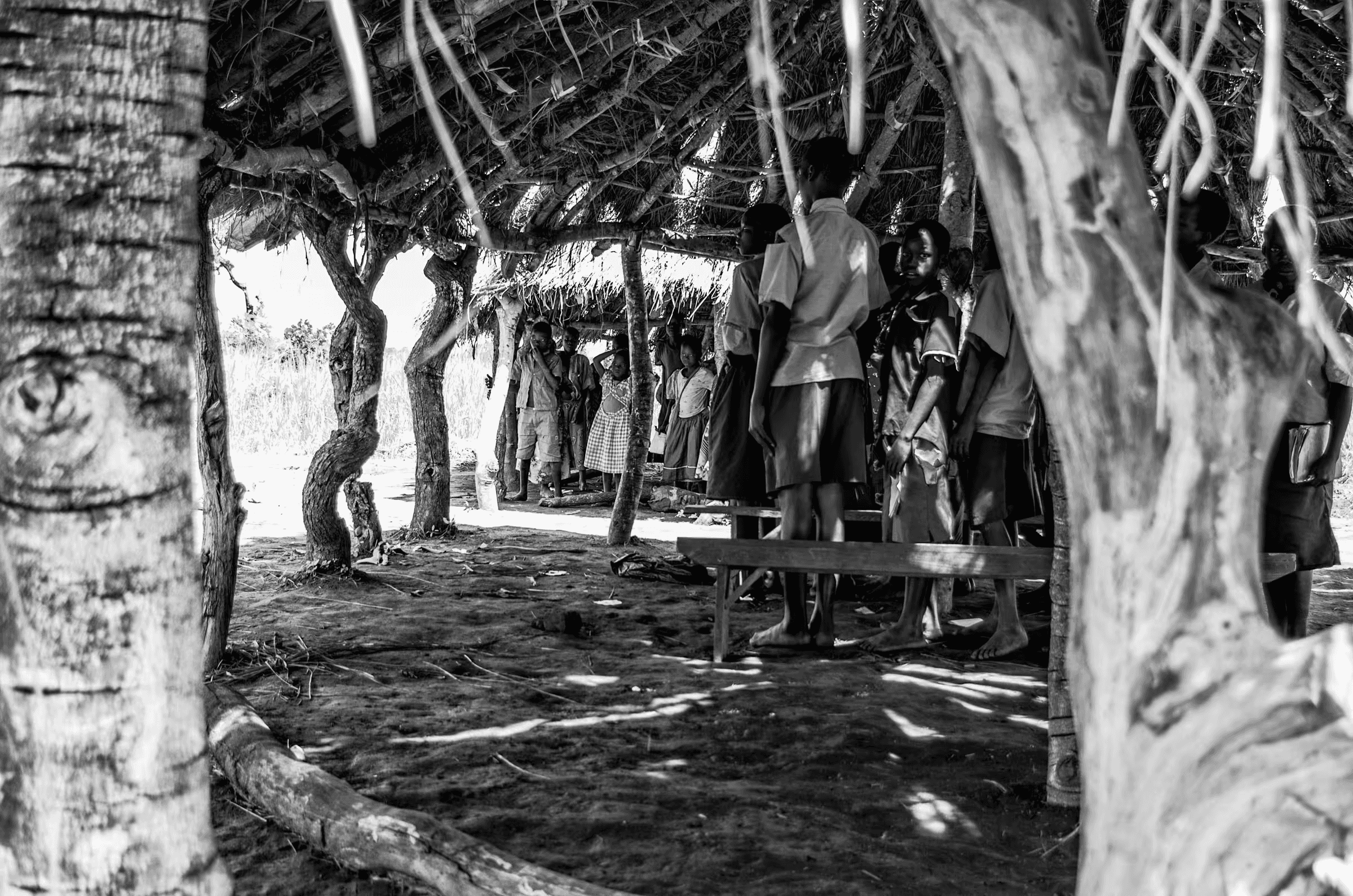 Black and white photograph showing a group of people, mostly children and some adults, gathered under a rustic thatched-roof structure in a rural Ugandan village.