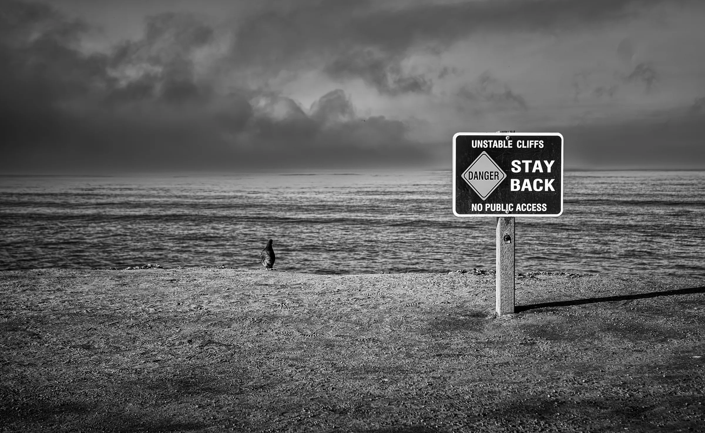 Black and white photograph of a warning sign reading 'Unstable Cliffs, Stay Back' on a beach, with the sea and a dramatic cloudy sky in the background.