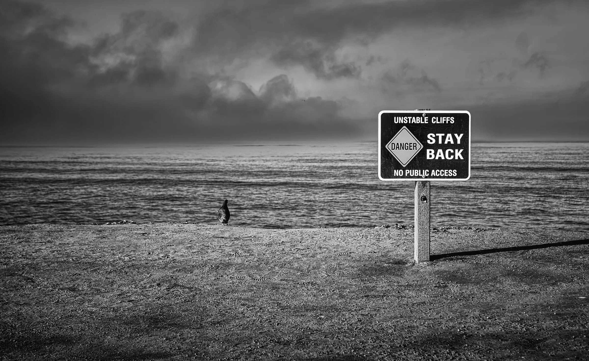 Black and white photograph of a warning sign reading 'Unstable Cliffs, Stay Back' on a beach, with the sea and a dramatic cloudy sky in the background.