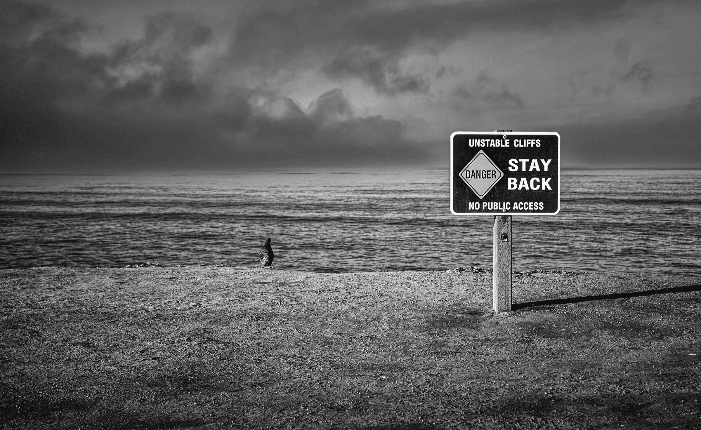 Black and white photograph of a warning sign reading 'Unstable Cliffs, Stay Back' on a beach, with the sea and a dramatic cloudy sky in the background.