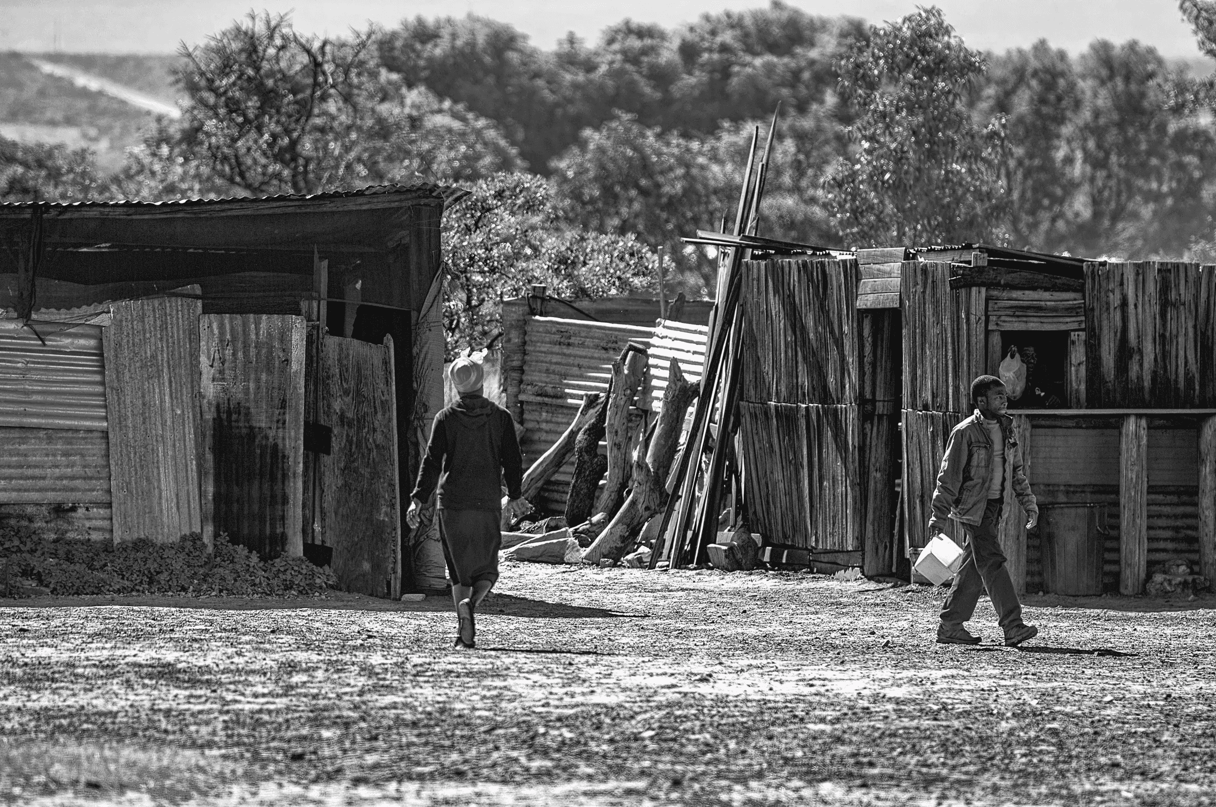 Black and white photograph showing two people walking between simple dwellings made of corrugated metal and wood in Vaalwater, South Africa.