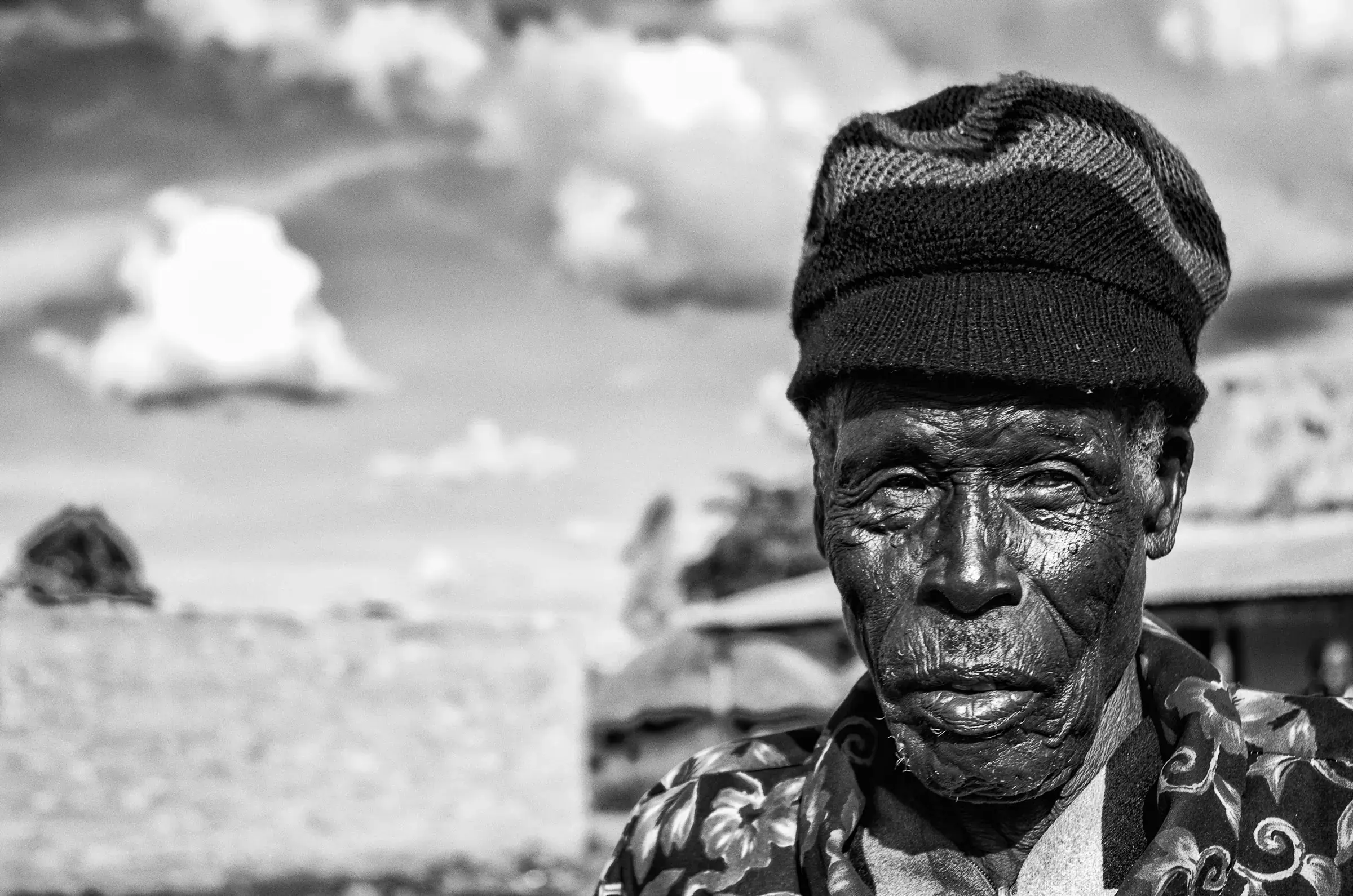 Weathered Gaze Black-and-white portrait of an older man in Uganda wearing a knit cap and patterned shirt, looking at the camera beneath a cloudy sky.