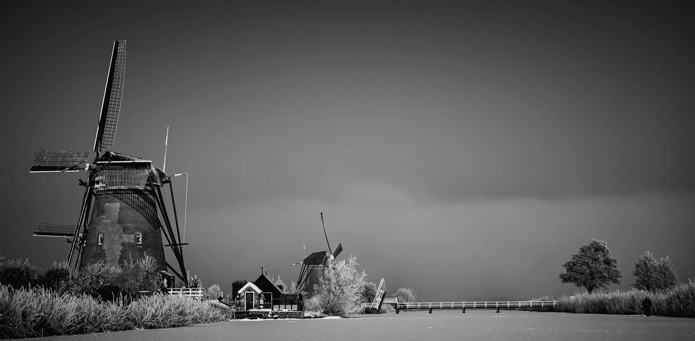 Black and white landscape photograph of windmills at Kinderdijk, Netherlands. Silhouetted windmills stand against a misty sky, with a small bridge and traditional Dutch architecture visible in the mid-ground.